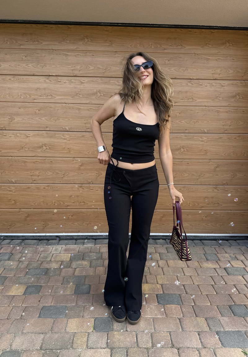 Donna con abbigliamento nero e occhiali da sole sorridente, che tiene una piccola borsa con stampa zebrata, in piedi su una superficie lastricata con muro di legno dietro.