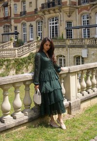 Vestido verde oscuro, con volantes, de mangas largas y escote en V. Los accesorios incluyen un pequeño bolso blanco y zapatos en tonos claros. Fondo de barandilla de piedra.