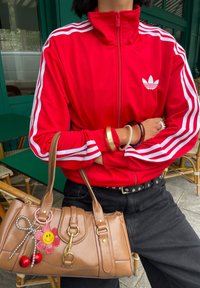 Asmeniui, vilkinčiam raudoną Adidas treninginę striukę su baltais dryžiais ir juodomis kelnėmis, rankoje laikanti rudą rankinę su šypseną simbolizuojančia gėle ir vyšnių raktų pakabuku.