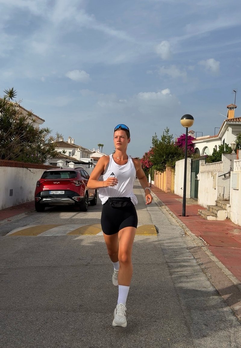 Jeune personne faisant du jogging dans une rue résidentielle, vêtue de vêtements de sport et de lunettes de soleil, avec des voitures garées et des maisons en arrière-plan.