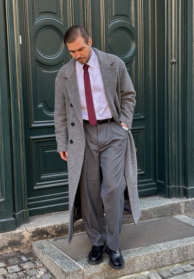 Hombre con abrigo largo gris, camisa blanca, corbata burdeos y pantalones grises, de pie sobre un escalón de piedra frente a una puerta verde oscuro.