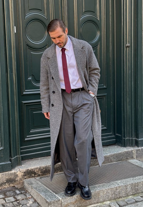 Mann in langem grauem Mantel, weißem Hemd, burgunderroter Krawatte und grauer Hose, stehend auf einem Steintritt vor einer dunkelgrünen Tür.