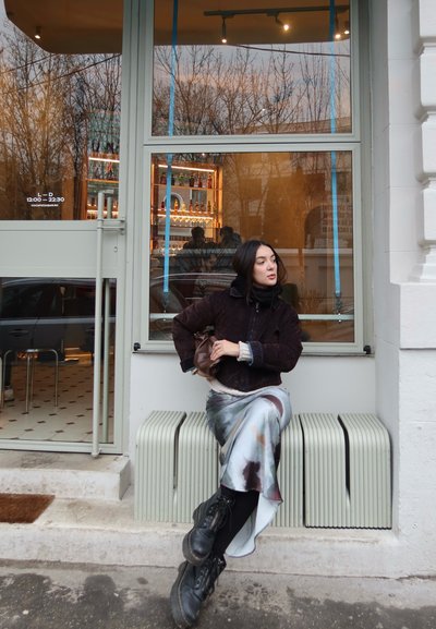 Mujer con chaqueta oscura, falda con estampado y botas sentada en un banco fuera de un café con una ventana de cristal que refleja árboles y botellas en el interior.