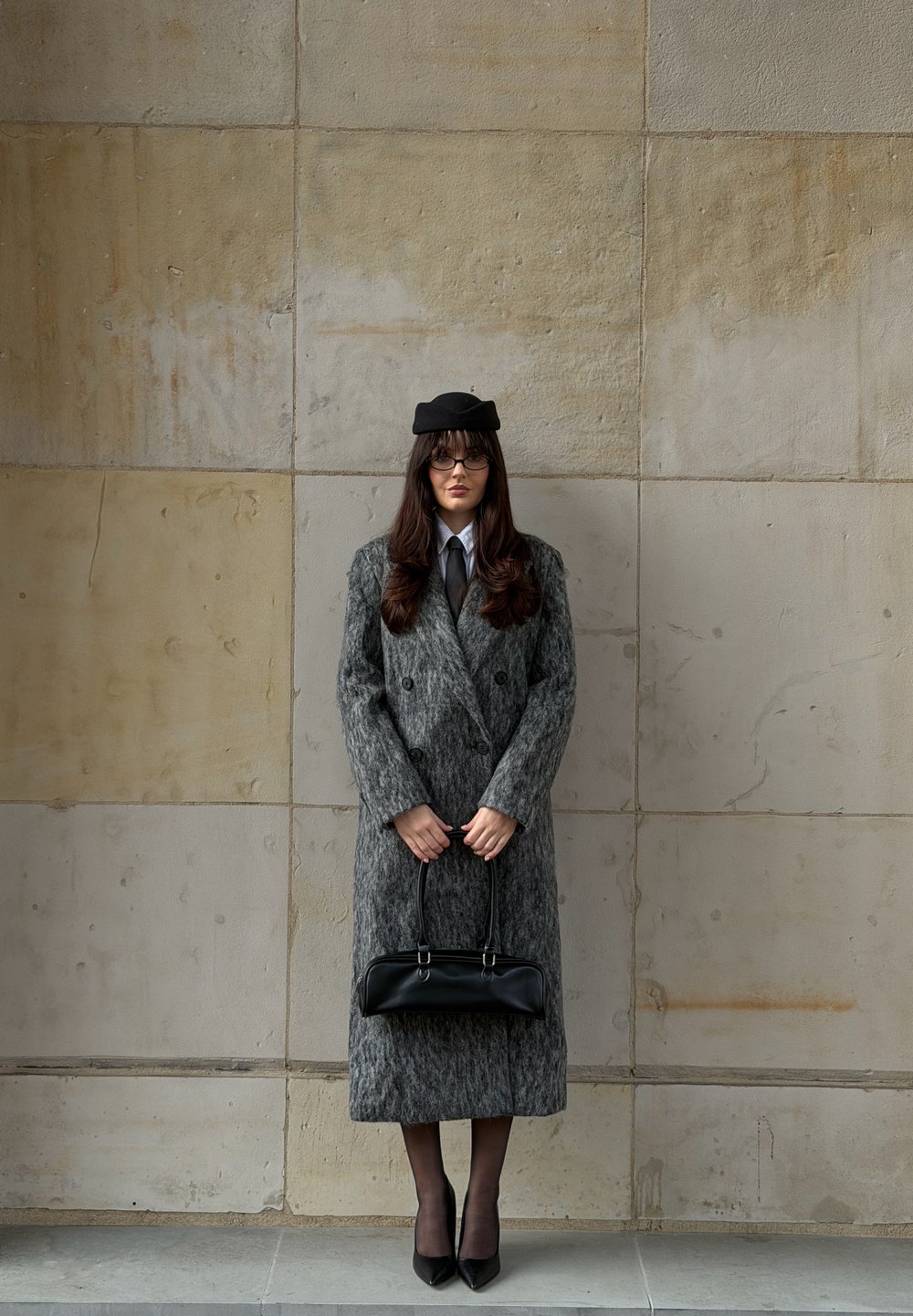 Manteau gris en laine à double boutonnage avec tissu texturé, sac à main noir et béret assorti. Escarpins noirs remarquables et chemise bleu clair.