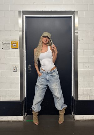 Top blanco sin mangas con cortes, jeans de mezclilla holgados de color azul claro, botas de tobillo beige sueltas y una gorra de color marrón claro, de pie frente a una puerta oscura.