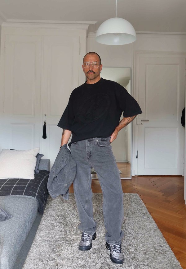 Homme avec lunettes et moustache se tient à l’intérieur tenant une veste grise, portant un t-shirt noir surdimensionné, un jean gris et des baskets sur un tapis.