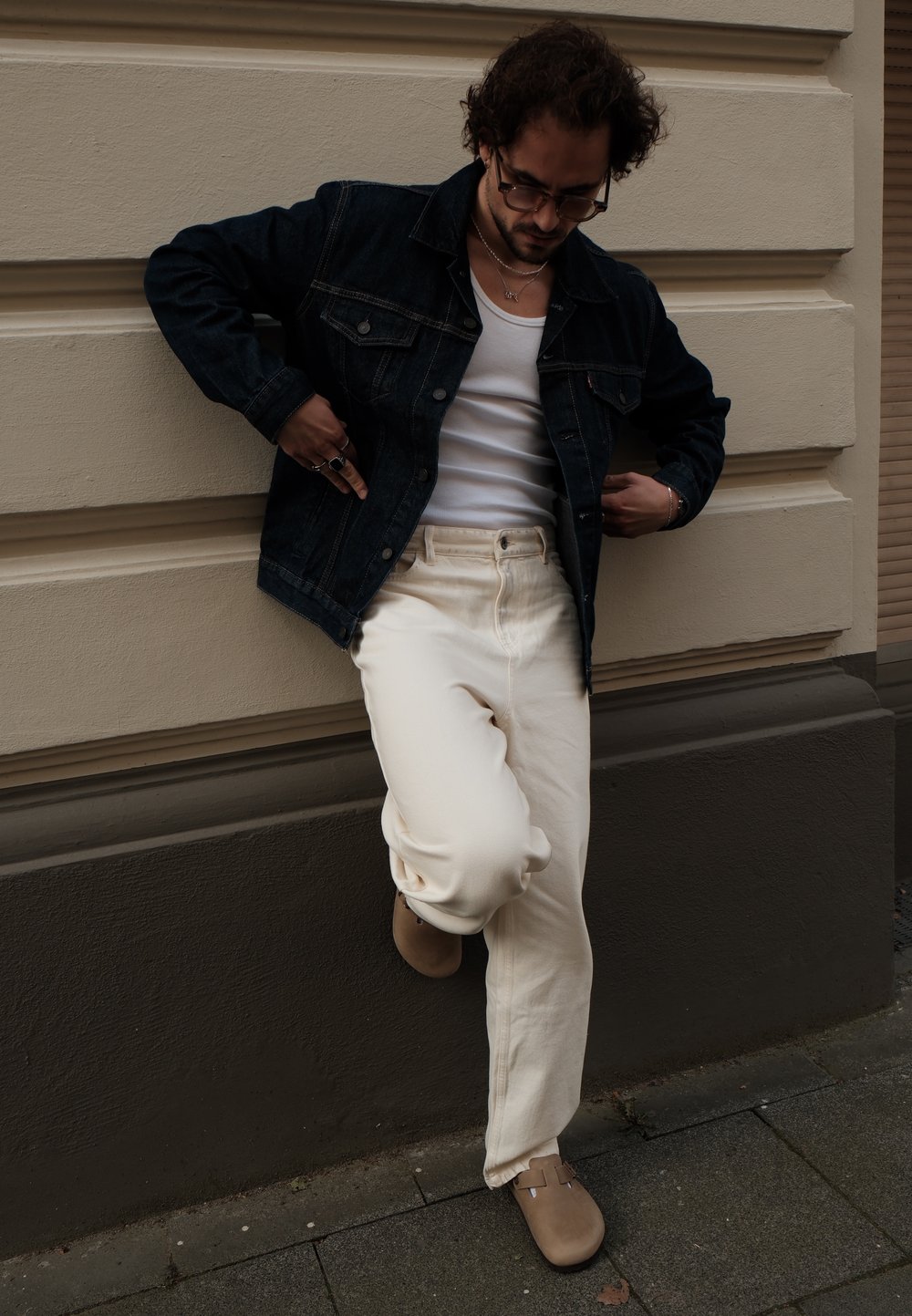 Giacca di jeans, canotta bianca, pantaloni larghi color crema, calzature marroni con fibbia. Gli accessori includono anelli e collane, sfondo di parete testurizzato.