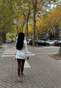 Nainen valkoisessa mekossa ja korkeakoroisissa koroissa kävelee puiden reunustamaa kaupunkikatujalkakäytävällä kohti suojatietä, edessä pysäköityjä autoja ja rakennuksia.