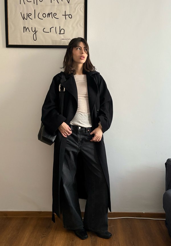 Giovane donna con cappotto lungo nero e jeans larghi in piedi vicino a un muro con testo incorniciato "Hello MTV welcome to my crib."