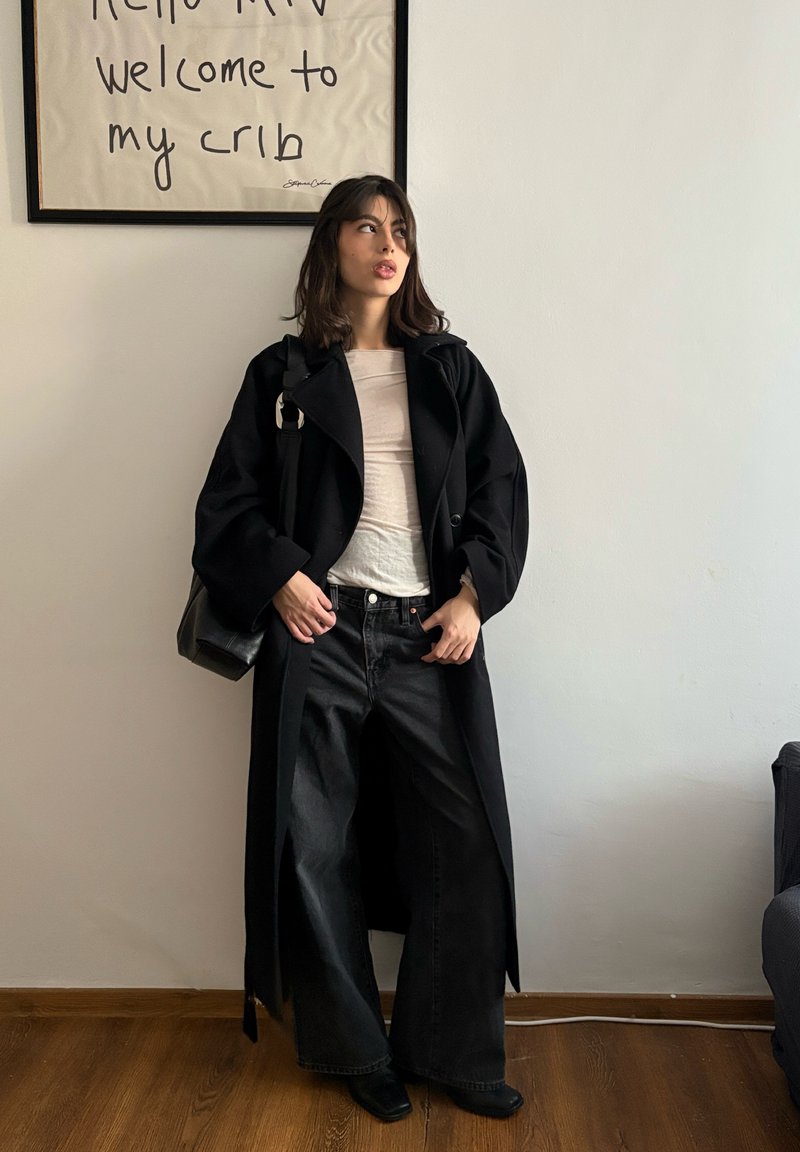 Jeune femme en manteau long noir et jeans larges debout près d'un mur avec un texte encadré "Hello MTV welcome to my crib."