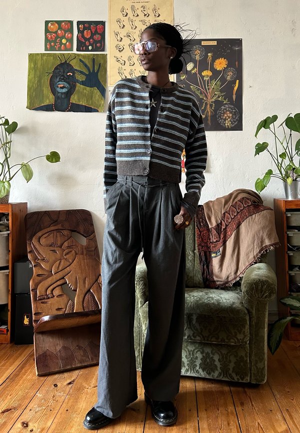 Femeie cu cardigan cu dungi și pantaloni largi stă într-o cameră confortabilă cu plante, fotoliu verde, sculptură din lemn în formă de elefant și postere botanice pe perete.
