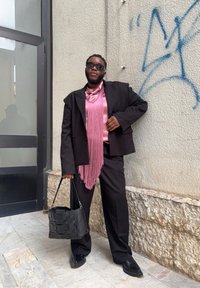 Čierny blazer nad ružovou blúzkou s třásňami, spárovaný s čiernymi nohavicami a tkanou čiernou taškou. Slnečné okuliare dopĺňajú look. Hladké textúry.