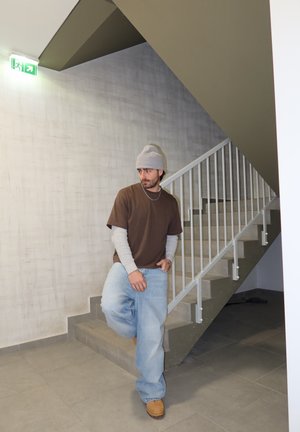 Joven con gorro, camisetas marrón y gris superpuestas, vaqueros holgados y botas, apoyado contra unas escaleras bajo un letrero de salida de emergencia.