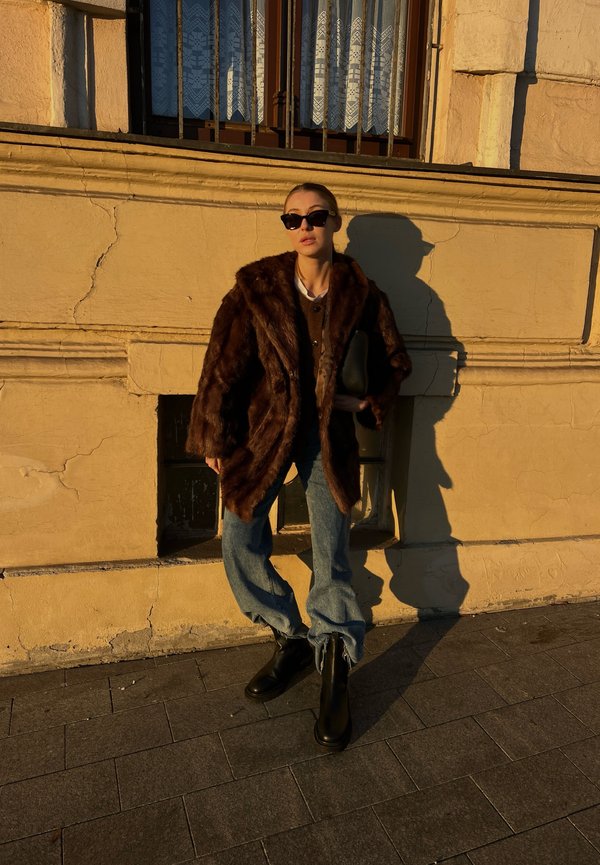 Brun fuskpälsjacka, oversized design, kombinerad med djupt blå avslappnade jeans och svarta chunky stövlar. Tillbehör inkluderar en svart kuvertväska och solglasögon.