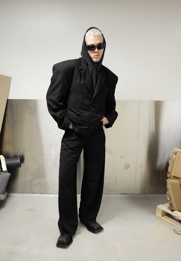 Osoba o platynowo blond włosach, ubrana w czarną, oversize'ową marynarkę, bluzę z kapturem, spodnie z szerokimi nogawkami i okulary przeciwsłoneczne, stojąca w przestrzeni przemysłowej.