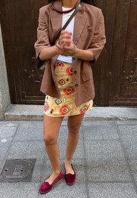 Brązowa lniana marynarka na białej bluzce, zestawiona z wzorzystą żółtą mini spódnicą z czerwonymi i niebieskimi akcentami oraz burgundowymi pantoflami wsuwanymi.