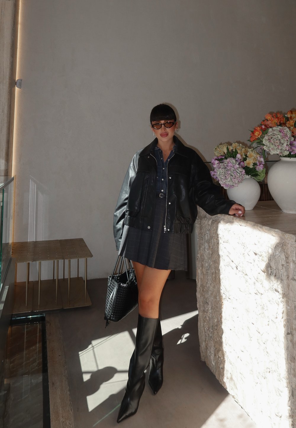 Chaqueta de cuero negra, camisa de mezclilla, falda de cuadros, botas negras hasta la rodilla, bolsa negra tejida, rodeada de arreglos florales en jarrones blancos.