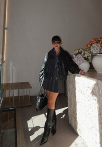 Chaqueta de cuero negra, camisa de mezclilla, falda de cuadros, botas negras hasta la rodilla, bolsa negra tejida, rodeada de arreglos florales en jarrones blancos.