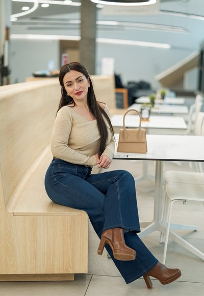 Mujer con cabello largo y oscuro sentada en un banco de madera en un café moderno, vistiendo un suéter beige, jeans azules, tacones marrones, con un bolso beige sobre la mesa.