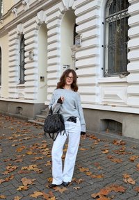 Pull gris tricoté avec accents en dentelle, associé à un pantalon blanc à jambes larges. Sac à main noir avec des anses. En arrière-plan, un bâtiment et des feuilles tombées.