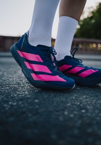 Blå sportskor med rosa diagonala ränder och snören, med en texturerad ovandel och en gummisula, placerade på en asfaltyta.