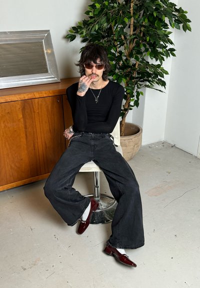 Camisa negra de manga larga, jeans negros de pierna ancha, zapatos color burdeos, cadena de plata, sentado en un taburete blanco junto a un armario de madera y una planta en maceta.