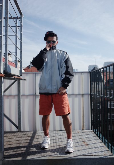 Chaqueta con cremallera en gris claro y negro, pantalones cortos acolchados naranjas, zapatillas blancas. Los accesorios incluyen gafas de sol y una gorra. Entorno urbano en una azotea.