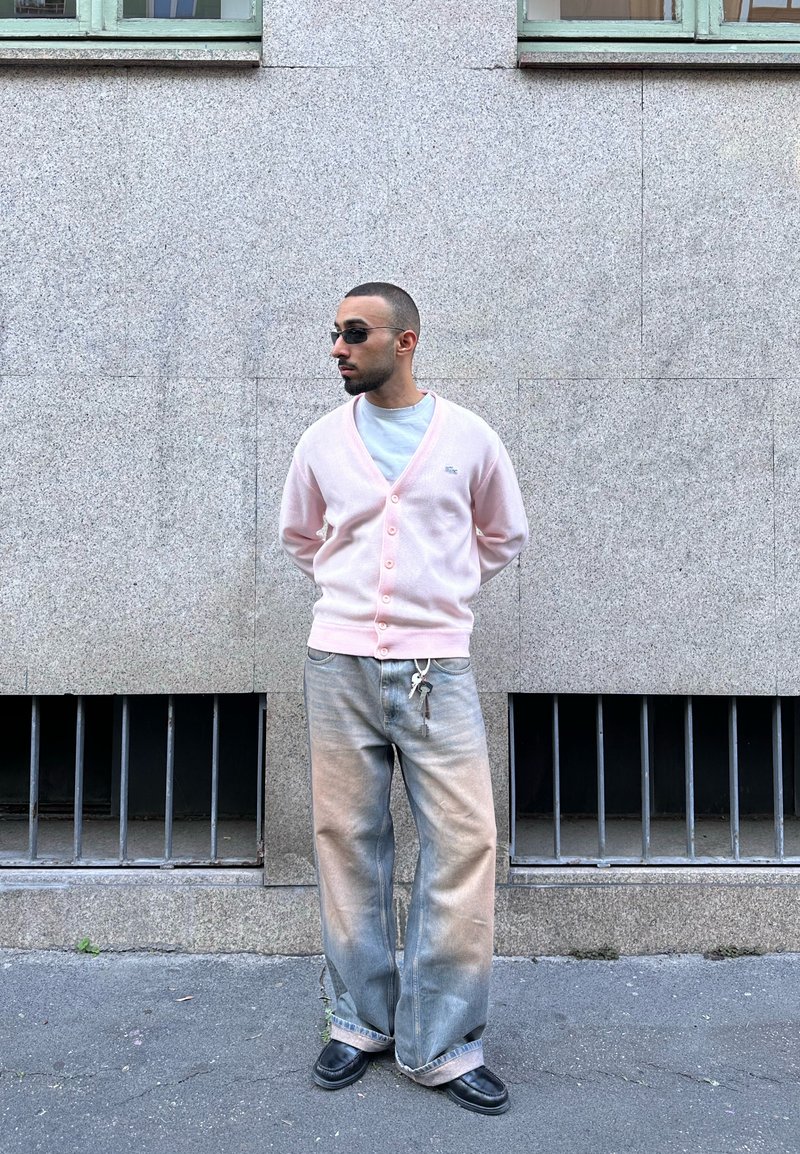 Homme portant un cardigan rose pâle, un jean gris à jambes larges avec des clés attachées, des chaussures noires et des lunettes de soleil, debout contre un mur en pierre avec des fenêtres barricadées.