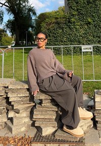Pullover grigio a maglia, pantaloni larghi scuri e scarpe beige. Seduto su blocchi di pietra sovrapposti in un ambiente verde all'aperto con alberi.