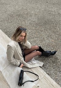 Bézs, dupla soros kabát szürke pulóver fölött, fekete miniszoknya és térdig érő fényes fekete csizmák. Mellette fekete oldaltáska. Texturált kavicsos háttér.