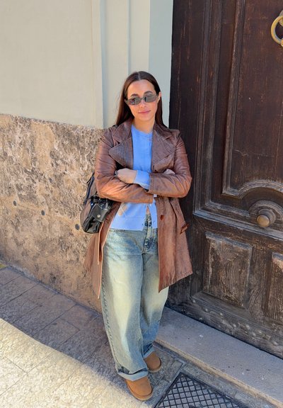 Mujer con chaqueta de cuero marrón, cárdigan azul, vaqueros y gafas de sol apoyada contra una pared cerca de una gran puerta de madera.