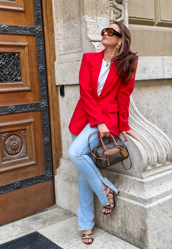 Una mujer con una americana roja, gafas de sol, vaqueros y sandalias posa junto a una puerta de madera ornamentada, sosteniendo un bolso.