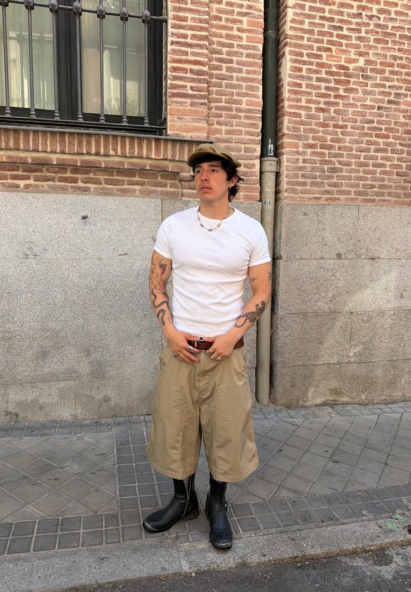 Bărbat cu tatuaje, purtând un tricou alb, pantaloni scurți bej până la genunchi, cizme negre și șapcă maro, stă pe trotuar lângă un zid de cărămidă și piatră.