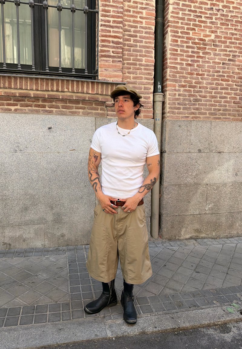 Homme avec des tatouages portant un t-shirt blanc, un short beige jusqu'aux genoux, des bottes noires et une casquette marron, debout sur le trottoir contre un mur en brique et pierre.