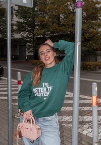 Zelená mikina s bílým textem, světle modré džíny a malá růžová kabelka se zlatými akcenty, stojící u kovového sloupu v městském prostředí.
