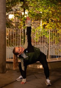 Moteris sportiniais drabužiais tempiasi lauke naktį, pakeldama vieną ranką į viršų ir šypsodamasi, o fone matyti rudens medžių ir turėklų.