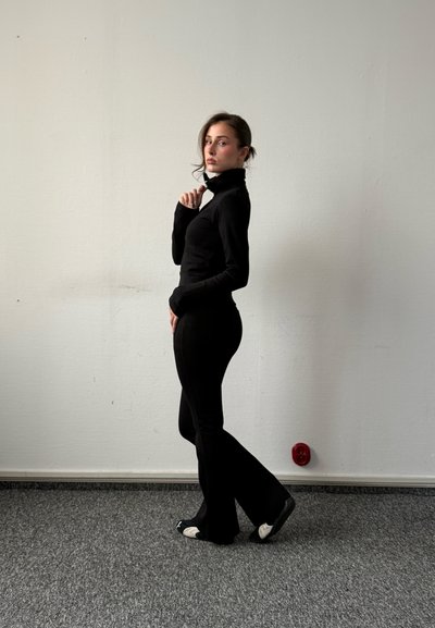 Mujer con jersey de cuello alto negro y pantalones, de pie de lado sobre suelo alfombrado gris con pared blanca de fondo, sujetando el cuello cerca de la barbilla.
