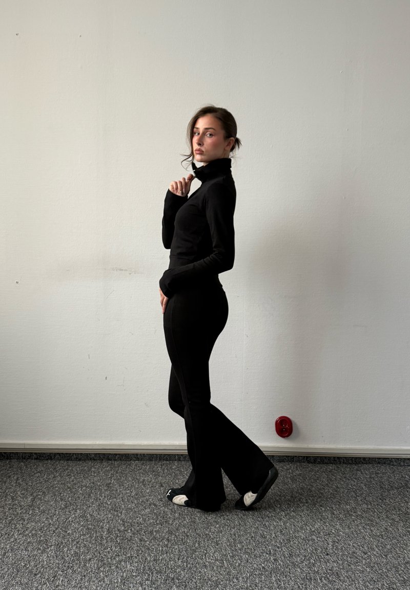 Femme en col roulé noir et pantalon, debout de profil sur un sol moquetté gris avec un mur blanc en arrière-plan, tenant le col près de son menton.