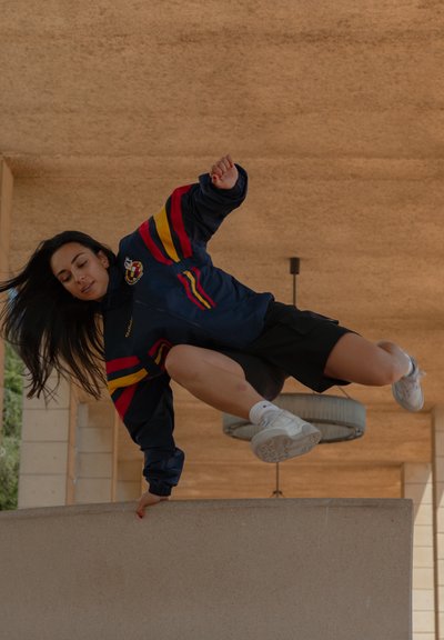 Una persona con una chaqueta azul con rayas rojas y amarillas y pantalones cortos negros salta sobre una barrera de concreto, llevando zapatillas blancas.