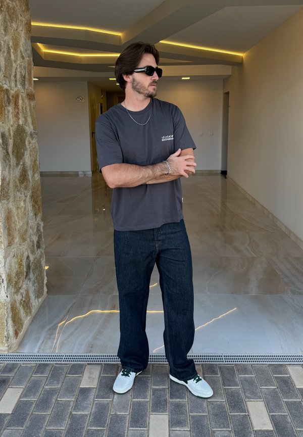 Grey short-sleeve t-shirt, dark blue loose-fitting jeans, white sneakers with green accents. Standing in a modern interior with stone walls.