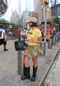 Svart håndveske med stjernedetalj, gul t-skjorte med grafikk, camo-mønstrets skjørt og svarte gummistøvler. Sidelengdesprofil i en urban setting.