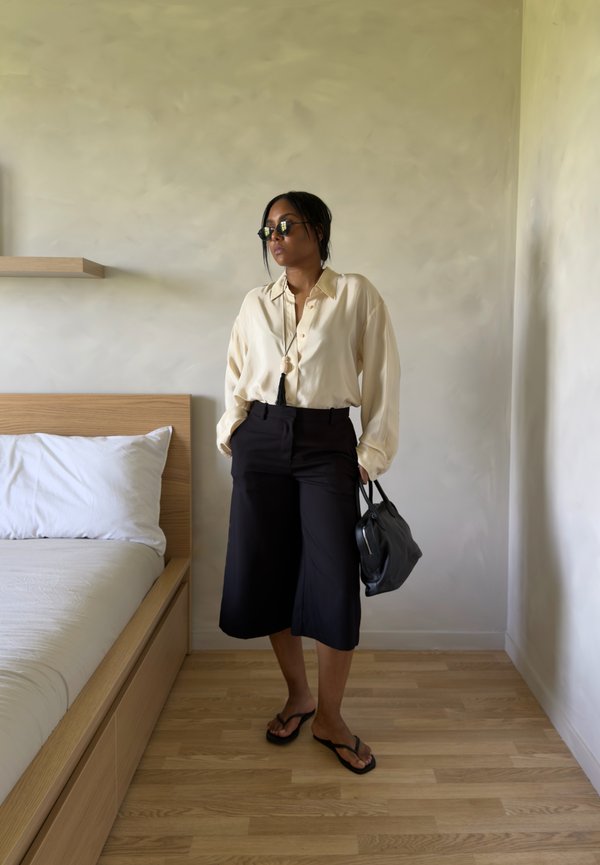 Femme portant des lunettes de soleil, blouse beige, short large noir, tenant un sac à main noir, debout à côté d'un lit dans une pièce minimaliste.