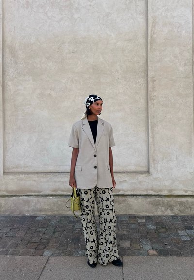 Blazer oversized de color beige claro con botones negros, combinado con pantalones de pierna ancha con un patrón floral en crema y negro. Lleva un pequeño bolso amarillo.