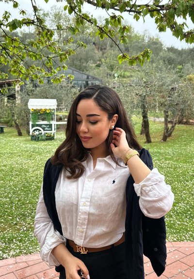 Camisa blanca de lino con cuello tipo button-down, suéter azul marino drapeado sobre los hombros, reloj de pulsera dorado, cinturón marrón con logo, en un entorno exterior verde.