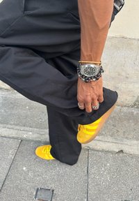 Osoba w czarnych spodniach podnosząca nogę, nosząca jasnobiałe trampki, srebrny zegarek oraz wiele bransoletek i pierścionków na lewej ręce, na tle betonu.
