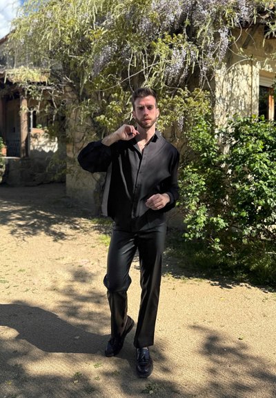 Camisa negra y transparente de manga larga con un corte relajado, combinada con pantalones negros ajustados y zapatos negros. Fondo natural con vegetación.