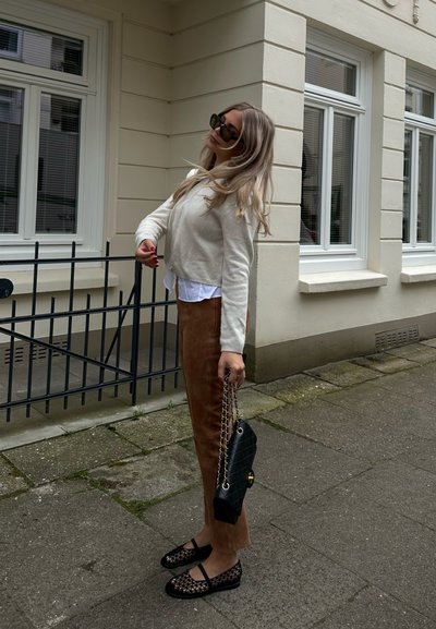 Un suéter crema sobre una camisa blanca, pantalones de ante marrón, un bolso negro acolchado con cadena, y bailarinas de malla negras, de pie sobre un pavimento de piedra.