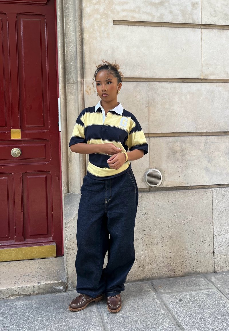 Jeune femme avec des cheveux bouclés en chignon, portant une chemise de rugby rayée jaune et noire et un jean noir ample, se tenant contre un mur en pierre près d'une porte rouge.