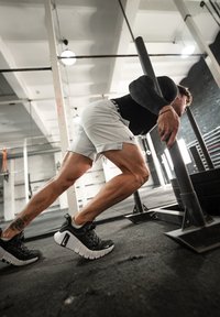 Sportlane, kes tõukab kaaluhaaki, seljas must pikka varrukat, hallid lühikesed püksid. Jalatseid on mustad valgete tallaga, tekstureeritud pinnaga.