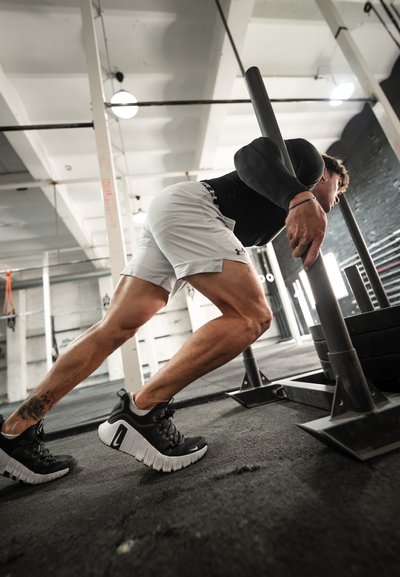 Atleta empujando un trineo con peso, vistiendo una camiseta de manga larga negra y pantalones cortos gris claro. El calzado es negro con suelas blancas y superficie texturizada.